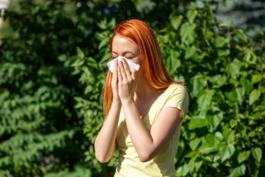 Genç kızıl saçlı kadın çiçek açan ağacın önünde hapşırıyor. Polen alerjisi belirtileri, arka planda yeşil park