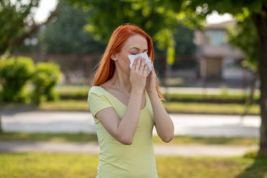 Genç kızıl saçlı kadın çiçek açan ağacın önünde hapşırıyor. Polen alerjisi belirtileri