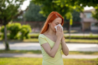 Genç kızıl saçlı kadın çiçek açan ağacın önünde hapşırıyor. Polen alerjisi belirtileri