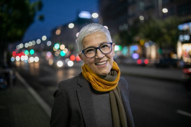 Gülümseyen, yaşlı, kısa gri saçlı ve gözlüklü yaşlı bir kadın sokakta yürüyor.