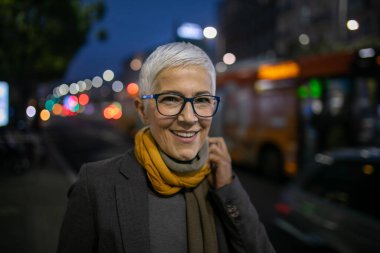 Gülümseyen, yaşlı, kısa gri saçlı ve gözlüklü yaşlı bir kadın sokakta yürüyor.