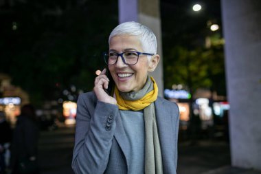 Gülümseyen, yaşlı, kısa gri saçlı, gözlüklü bir kadın. Sokakta, şehir merkezinde, gece görüş alanında akıllı telefon kullanıyor.