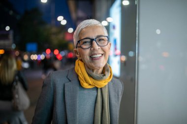 Gülümseyen, yaşlı, kısa gri saçlı ve gözlüklü yaşlı bir kadın. Sokakta yürüyor.