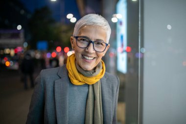 Gülümseyen, yaşlı, kısa gri saçlı ve gözlüklü yaşlı bir kadın. Sokakta yürüyor.