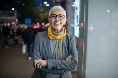 Gülümseyen, yaşlı, kısa gri saçlı ve gözlüklü yaşlı bir kadın. Sokakta yürüyor.