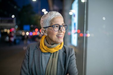 Gülümseyen, yaşlı, kısa gri saçlı ve gözlüklü yaşlı bir kadın. Sokakta yürüyor.