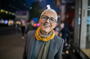 Gülümseyen, yaşlı, kısa gri saçlı ve gözlüklü yaşlı bir kadın. Sokakta yürüyor.