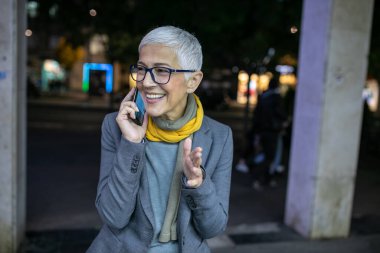 Gülümseyen, yaşlı, kısa gri saçlı, gözlüklü bir kadın. Sokakta, şehir merkezinde, gece görüş alanında akıllı telefon kullanıyor.