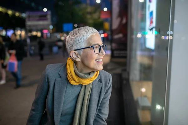 Smiling mature senior woman with short gray hair and eyeglasses looking in the shop window, city night scene