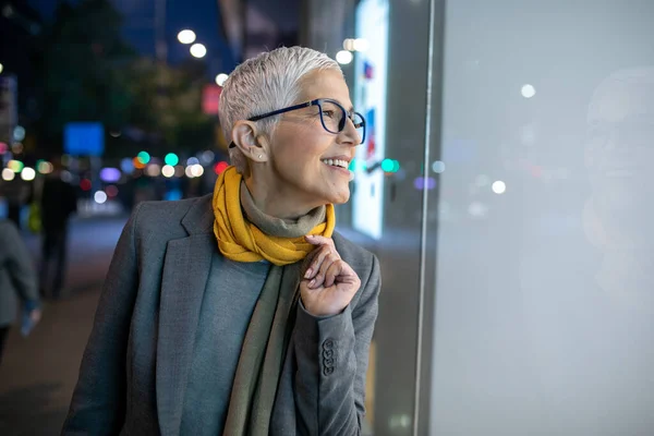 Smiling mature senior woman with short gray hair and eyeglasses looking in the shop window, city night scene