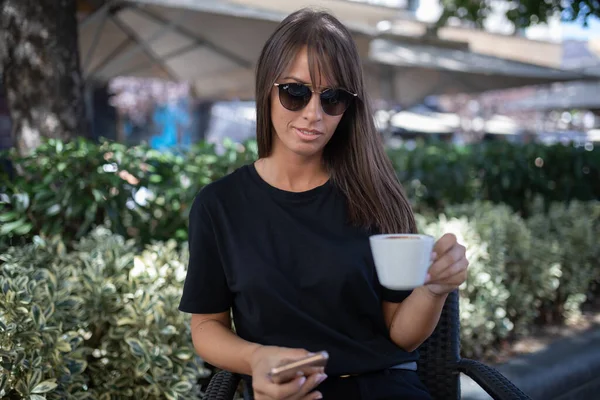 カフェでの朝 黒のドレスとダークサングラスの魅力的な女性はコーヒーを飲み ソーシャルネットワークのための自撮り写真を作ります ストックフォト — ストック写真