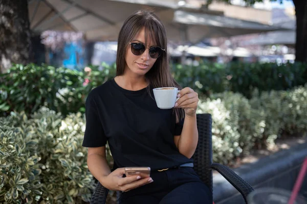 カフェでの朝 黒のドレスとダークサングラスの魅力的な女性はコーヒーを飲み ソーシャルネットワークのための自撮り写真を作ります ストックフォト — ストック写真