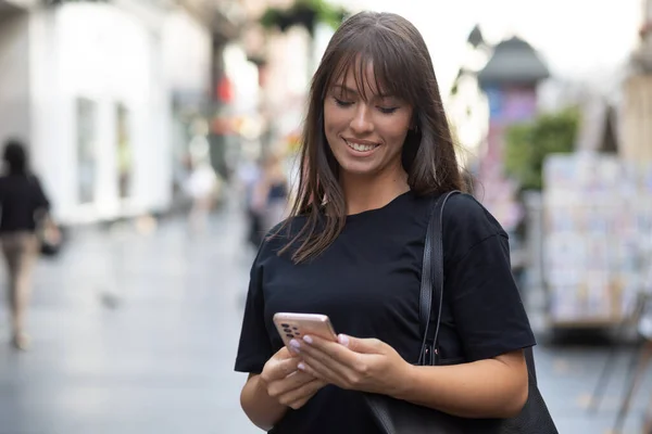 美しい笑顔の女性で黒シャツのテキストメッセージスマートフォンの路上で市内中心部 — ストック写真