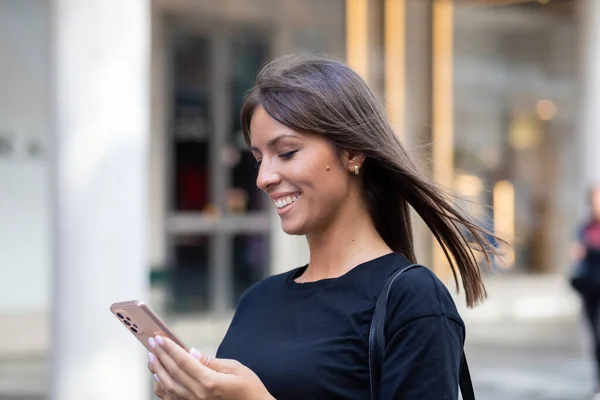 Beautiful smiling woman in black shirt texting on smartphone on street in city center