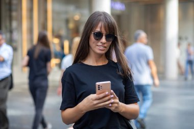 Siyah gömlekli ve güneş gözlüklü güzel bir kadın şehir merkezinde akıllı telefondan mesaj atıyor.