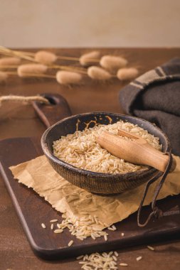 Brown rice in ceramic bowl with wooden scoop on rustic countertop.