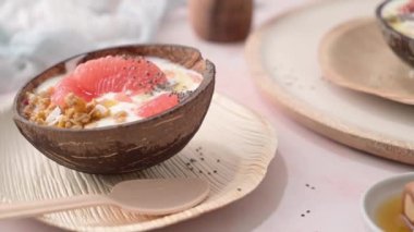 Yogurt with grapefruit, granola, chia and honey served in half a coconut shell on a rose marble kitchen countertop.
