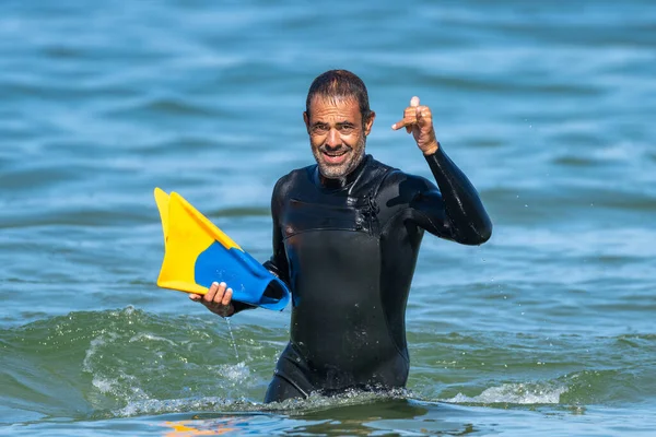 Middle-aged dark-haired man with sea water up to his waist dressed in a thermal suit, with fins in one hand and greeting with the other.