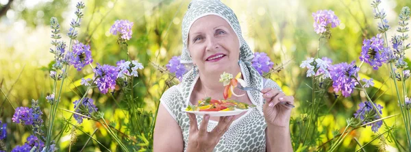 Uma mulher idosa come salada de legumes no fundo de flores de prado — Fotografia de Stock
