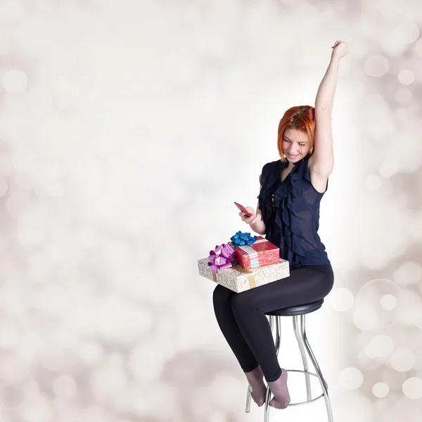 Happy red-haired girl with gifts and phone on a chair at a festive abstract background — Stock Photo, Image