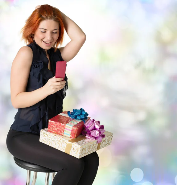 Mujer pelirroja feliz con una caja de regalo y un teléfono en el fondo festivo abstracto —  Fotos de Stock