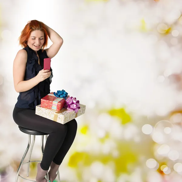 Donna dai capelli rossi felice con una scatola regalo e un telefono sullo sfondo festivo astratto — Foto Stock