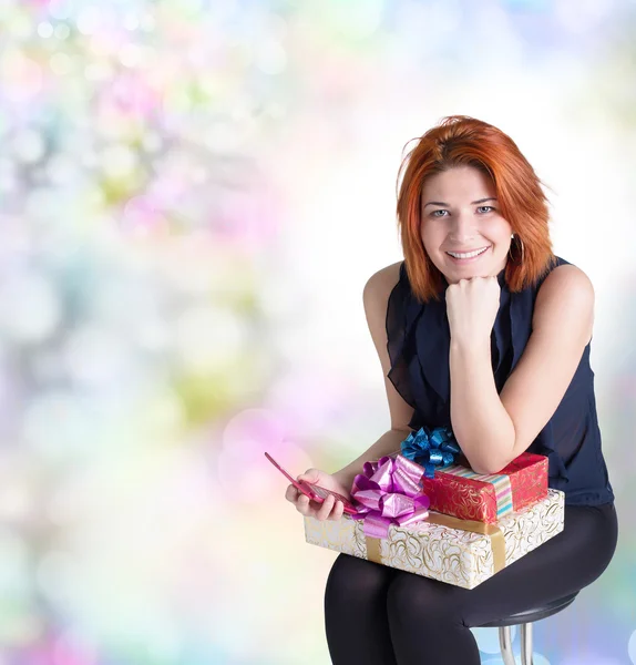 Mujer pelirroja feliz con una caja de regalo y un teléfono en el fondo festivo abstracto —  Fotos de Stock