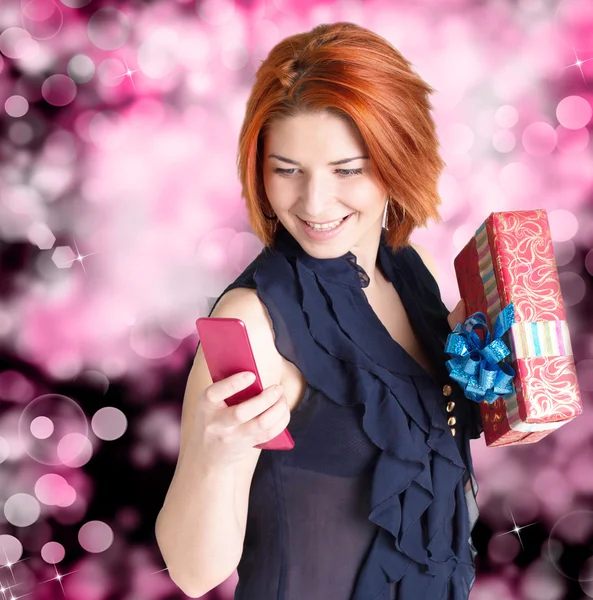 Femme aux cheveux roux souriante avec des boîtes cadeaux et téléphone sur un fond de fête — Photo