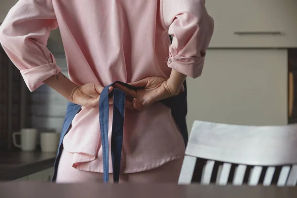 Mujer en traje rosa ata delantal azul en su espalda para empezar a cocinar en su cocina casera. Imagen sin rostro. — Foto de Stock