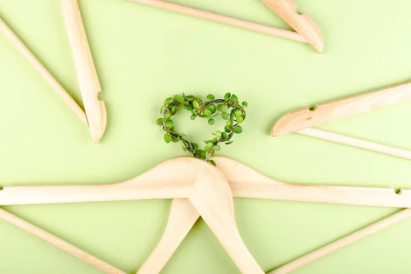 Conscious consumption slow fashion concept. Heart of clothes hangers entwined with plant on green background.