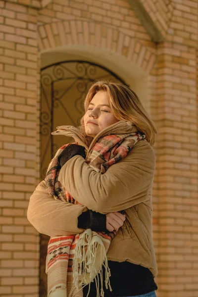 Mujer joven rubia en chaqueta beige y bufanda se envolvió contra el fondo del edificio de ladrillo. Temporada de invierno. De cerca. —  Fotos de Stock