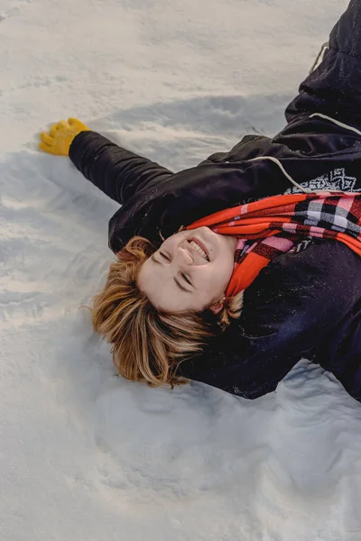 Feliz joven rubia yace en la nieve con abrigo negro, bufanda naranja y guantes amarillos. Temporada de invierno. De cerca. —  Fotos de Stock