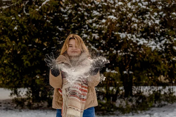 Blond ung kvinna i beige jacka och halsduk kastar snö med händerna. Bakgrund av julgranar i snö. Vintersäsong. — Stockfoto