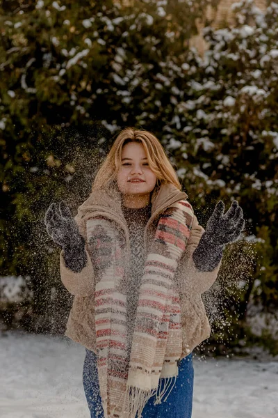 穿着米色夹克和围巾的金发年轻女子用手扔着雪。白雪中的圣诞树的背景。冬季. — 图库照片