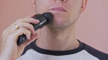 Genç adam içeride pembe bir arka planda elektrikli tıraş makinesiyle sakalını tıraş ediyor. Ön görünüm sabit yüksek kalite 4K görüntü