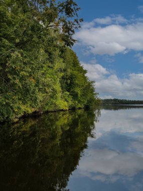 Karmaşık ormanlarla kaplanmış sarp kıyı gölün suyuna yansıyor. Güneşli bir yaz günü. Beyaz bulutlu mavi gökyüzü suya yansıyor..