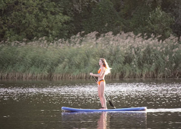 Sup surfing, happy blonde girl with paddle sailing on a board in river. Stand up paddle boarding in summer