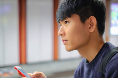 Young handsome asian man looking forward while using cellphone checking social media, surfing internet, and mobile apps at weekend at subway