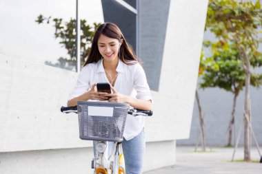 Kulaklıklı gülümseyen kız, elinde akıllı telefon, parkta bisiklet sürüyor.