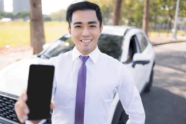 黒いスクリーンを持った携帯電話を見せ車の前に立つ若い笑顔のビジネスマン — ストック写真