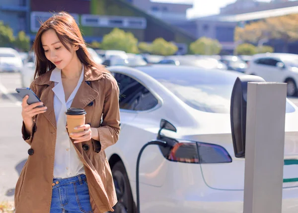 電気自動車の近くの駐車場に立つ若い女性の笑顔 小さな駅から自動車のバッテリーを充電し コーヒーを飲み スマートフォンを使用して — ストック写真