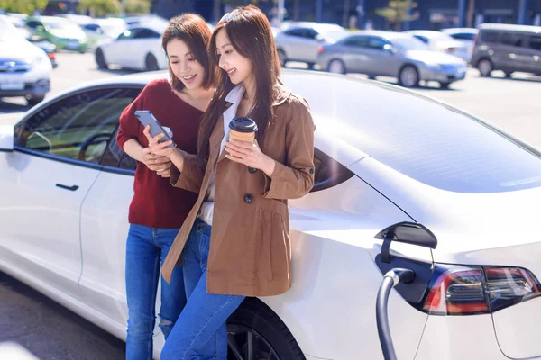電気自動車の近くの市内駐車場に立つ幸せな若い女性 小さな市内駅から自動車用バッテリーを充電し スマートフォンを見て情報を確認する — ストック写真