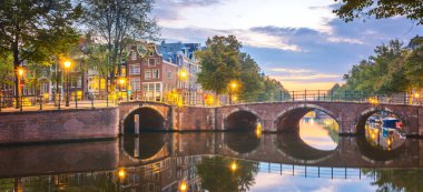 Büyülü Amsterdam. Şafak vakti Amsterdam 'ın panoramik manzarası. Şehir merkezinde. Fenerler hala açık, ama güneş çoktan gökyüzünü çok renkli renklere boyadı. Resimli, sessiz bir sabah. Sudaki yansımalar. Köprüler, kanallar ve evler. Avrupa