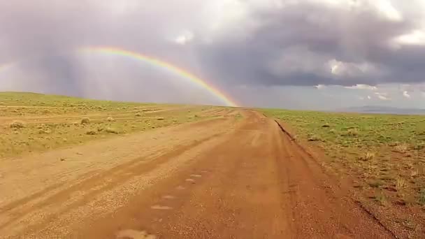 Deserto dirigindo em uma estrada lamacenta com um arco-íris na parte de trás — Vídeo de Stock