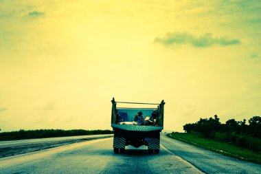 Two people traveling in back of a truck on an empty road in Cuba clipart