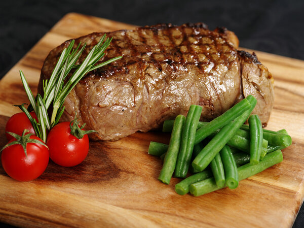 Photo of a thick sirloin steak dinner with rosemary, cherry tomatoes and green beans on a wooden board.