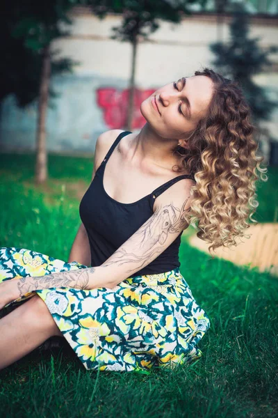 Şehir parkında çimlerde oturan kıvırcık saçlı kadının portresi.