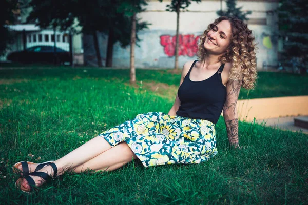 Şehir parkında çimlerde oturan kıvırcık saçlı kadının portresi.