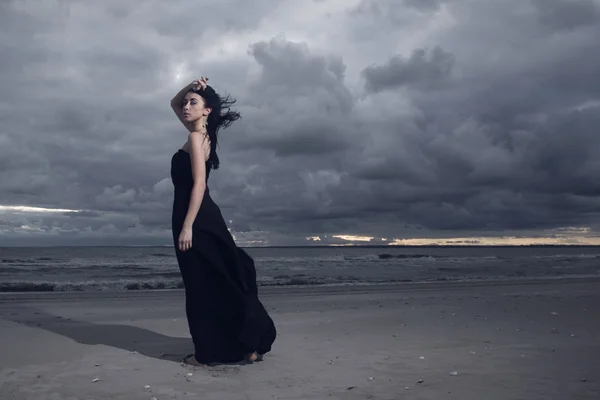 Magnifique modèle brune en robe longue noire debout sur la plage. Temps nuageux. Coucher de soleil — Photo