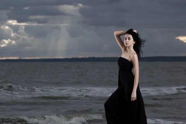 Gyönyörű barna modell a hosszú fekete ruhában állt a tengerparton. Felhős időjárás. Naplemente — Stock Fotó